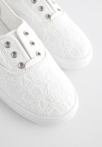 Baskets en toile blanche avec un motif brodé texturé, des œillets argentés et une semelle en caoutchouc. Bout arrondi et design discret.