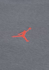 Γκρι ύφασμα με υφή, διακοσμημένο με κεντημένο κόκκινο λογότυπο Jumpman, που απεικονίζει τη σιλουέτα ενός παίκτη μπάσκετ σε κίνηση.