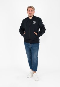 Giacca nera con zip e colletto tondo, caratterizzata da un logo sul lato sinistro del petto, abbinata a jeans in denim blu e sneakers bianche.