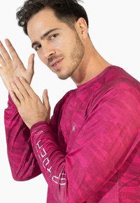 Pitkähihainen paita kirkkaan vaaleanpunaisena, jossa on teksturoitu kuvio ja logo hihassa. Kangas vaikuttaa kevyeltä ja joustavalta.