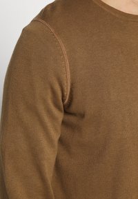 Brun stickad tröja med rund halsringning, med en mjuk textur och kontrasterande sömmar längs axeln.