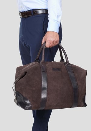 Brun semsket skinn duffelbag med mørke lærdetaljer, glidelås, og doble topphåndtak; har en rektangulær form og logo detalj.
