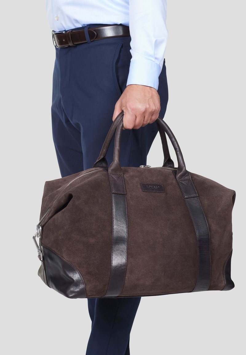 Brun semsket skinn duffelbag med mørke lærdetaljer, glidelås, og doble topphåndtak; har en rektangulær form og logo detalj.