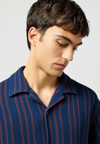 Gebreid blauwe shirt met verticale rode strepen, een kraag en knopen. De textuur is wafelachtig, met een comfortabele pasvorm en casual ontwerp.