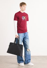 Burgundy pamut póló "Calvin Klein" logóval, fehér hosszúujjú ingre rétegzett. Hasított világoskék farmernadrág és fekete vászontáska.