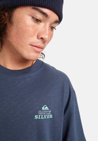 Mladý člověk v modrém čepici a modrém tričku Quiksilver se dívá dolů na pozadí čistě bílé stěny.