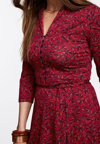 Rotes Blumenkleid mit Knopfleiste, Dreiviertelärmeln und einem taillierten Bund. Es verfügt über ein blätterförmiges Muster in Blau, Gelb und Grün.