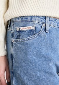 Detailní záběr na modré džínové kalhoty s etiketou Calvin Klein na malém kapsičce, nošené s krémovým pleteným svetrem a viditelnou rukou na boku.