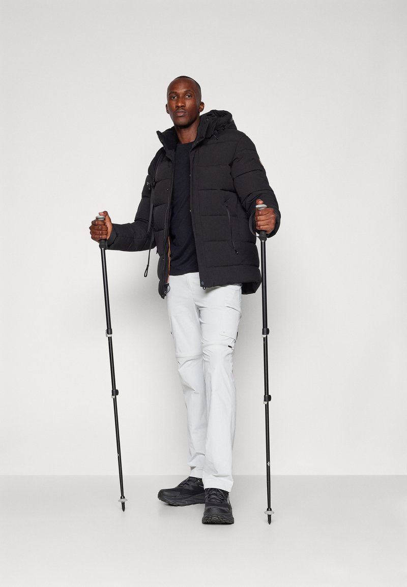 Schwarze gepolsterte Jacke mit Kapuze, kombiniert mit einem schwarzen Oberteil und hellen Hosen. Die Person hält Trekkingstöcke und trägt dunkle Schuhe.