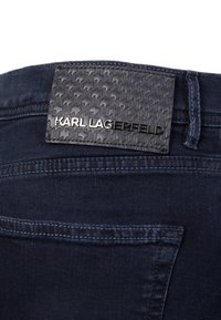 Black leather patch with embossed pattern and silver "KARL LAGERFELD" logo on the waistband of dark blue denim jeans.