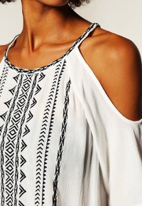 Vit kallskuldra topp med invecklad svart geometrisk broderi och ett mjukt, texturerat tyg. Justerbara remmar för en anpassningsbar passform.