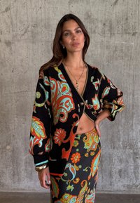 Γυναίκα που φοράει μαύρη ζακέτα και φούστα με πολύχρωμα πορτοκαλί, τυρκουάζ και πράσινα λουλουδάτα μοτίβα πεϊσλί, στέκεται μπροστά σε έναν τοίχο από σκυρόδεμα.