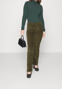 Žena ve tmavozeleném roláku, olivově zelených kordových kalhotách, černých podpatcích, držící malou černou kabelku, stojící na jednolitém pozadí.