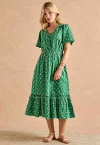 Zelené midi šaty s potlačou, krátkymi rukávmi, výstrihom do V, elastickým pásom a viacvrstvovým volánom na spodnej časti. Nosia sa s plochými sandálmi na remienky.