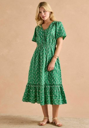 Grün gemustertes Midikleid mit kurzen Ärmeln, V-Ausschnitt, elastischer Taille und gestuftem Rüschensaum. Getragen mit flachen, geschnürten Sandalen.