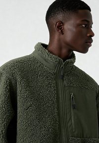 Giacca in pile verde oliva con superficie morbida e strutturata. Presenta una chiusura frontale con zip, colletto alto e una tasca sul petto con zip.