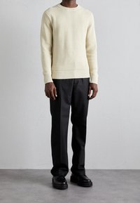 Cremfarbener, strukturierter Strickpullover mit gerippten Bündchen und Saum, kombiniert mit schwarzen, maßgeschneiderten Hosen und schwarzen, klobigen Schuhen. Ein schlichtes, modernes Outfit.