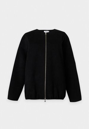 Femme portant une veste noire ample zippée, un pantalon noir droit et des chaussures noires, debout sur un fond clair uni.