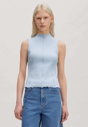 Jonge vrouw met lang blond haar, gekleed in een mouwloos lichtblauw geribd topje en een hoge taille blauwe spijkerbroek tegen een neutrale achtergrond.