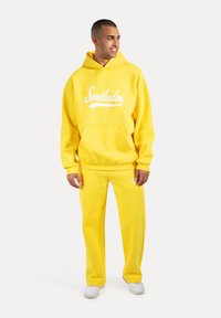 Żółta bawełniana bluza z kapturem i kieszenią kangurką, w komplecie z dopasowanymi żółtymi spodniami i białymi sneakerami.