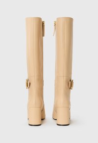Bottes en cuir beige jusqu'au genou avec des talons carrés, des accents en métal doré et une fermeture éclair sur le côté pour un enfilage facile. Texture lisse et design ajusté.