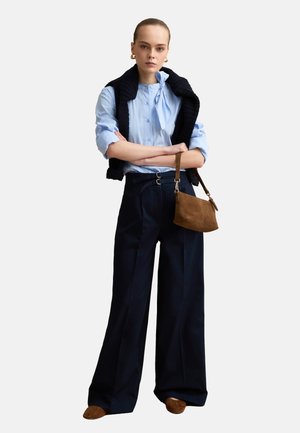 Frau trägt hellblaue Bluse mit Schleife, dunkelblaue weite Hose, schwarzen Strickpullover über die Schultern gelegt, hält braune Wildlederhandtasche.