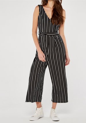 Femme portant une combinaison sans manches noire avec des rayures blanches verticales, une taille ceinturée, des jambes larges et des baskets blanches montantes.