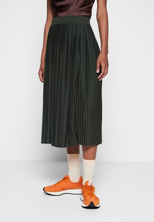 Kvinde iført mørkegrønt plisseret midinederdel, cremefarvede sokker og klare orange sneakers, stående mod en helt hvid baggrund.