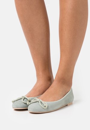 Ballerines en daim vert clair avec un nœud décoratif en corde torsadée, portées pieds nus sur un fond blanc.
