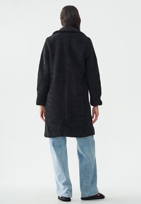 Abrigo negro de textura suave con cuello, mangas largas y corte recto, llevado sobre unos jeans de pierna ancha de color azul claro y calzado negro.