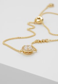 Collar en tono dorado con una gema redonda transparente, rodeada de piedras pequeñas transparentes. En una fina cadena con un bead deslizante para ajustar la longitud.