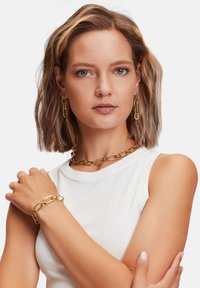 Set di gioielli in tonalità oro composto da una collana con catena spessa, bracciale coordinato e orecchini con design a maglie ovali indossati da un modello.