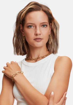 Ensemble de bijoux dorés comprenant un collier en chaîne épaisse, un bracelet assorti et des boucles d'oreilles au design en maillons ovales, porté par un modèle.