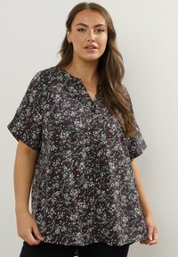 Donna che indossa una blusa nera a maniche corte con motivi floreali bianchi e viola, in piedi davanti a uno sfondo chiaro e uniforme.