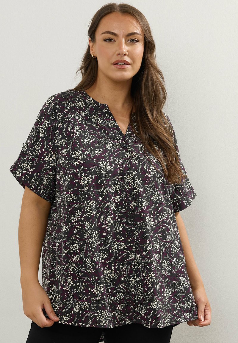 Donna che indossa una blusa nera a maniche corte con motivi floreali bianchi e viola, in piedi davanti a uno sfondo chiaro e uniforme.