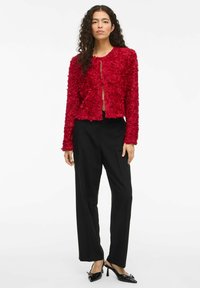 Veste rouge courte en tissu texturé avec des appliqués floraux, associée à un pantalon noir taille haute et des chaussures à talons noirs.