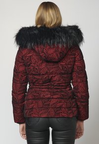 Kobieta w czerwoną wzorzystą kurtkę z kapturem wykończonym czarnym futrem, pokazana z tyłu, w połączeniu z czarnymi skórzanymi spodniami.