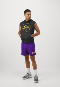Hoodie senza maniche nera con il logo "Lakers" in giallo e pantaloni corti in rete viola con il testo "Lakers". Scarpe da ginnastica grigie e calzini bianchi.