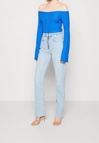 Blue ribbed off-the-shoulder top paired with light blue high-waisted jeans featuring a zipper. Model wears beige heeled sandals.