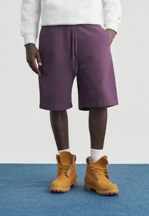 Άτομο με μαύρο δέρμα που στέκεται, φοράει μωβ σορτς, λευκές κάλτσες, καφέ εργασιακές μπότες και ένα ανοιχτό γκρι μακρυμάνικο πουκάμισο που φαίνεται μερικώς.