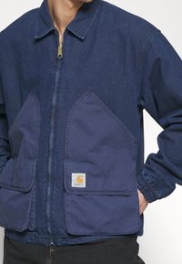 Veste en denim bleu avec une fermeture éclair, dotée de grandes poches avant pointues, de poignets côtelés et d'une étiquette de marque visible sur la poche.