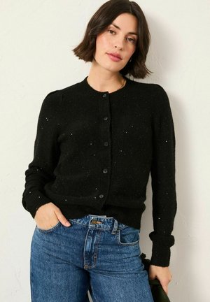 Schwarze gestrickte Strickjacke, runder Ausschnitt, lange Ärmel, mit kleinen Knöpfen und einem strukturierten Muster mit feinen Sprenkeln. Kombiniert mit hochgeschnittenen Jeans.