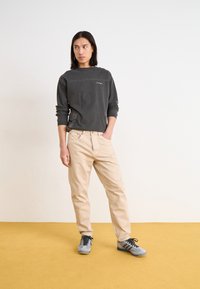 Haut gris à manches longues avec un petit graphique, associé à un pantalon beige clair. Les chaussures présentent des accents gris et noir. Debout sur un sol jaune.