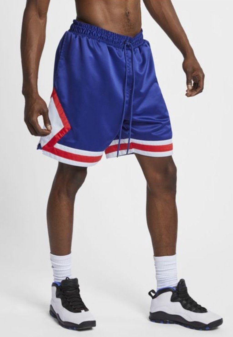 Homme portant un short de basket bleu avec des rayures rouges et blanches, des chaussettes blanches montantes et des chaussures de sport blanches et noires, debout sur fond blanc.