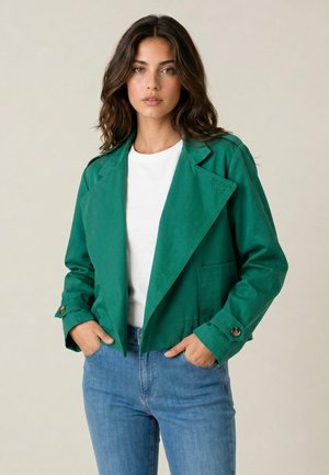 Donna con capelli scuri ondulati indossa una giacca corta verde sopra una camicia bianca e jeans blu, in piedi con le mani nelle tasche su uno sfondo uniforme.