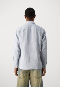 Persona vistiendo una camiseta de manga larga de color azul claro y jeans azules descoloridos, de pie de espaldas contra un fondo blanco liso.