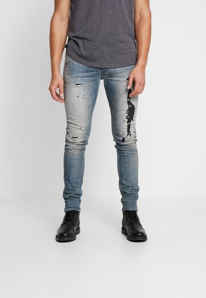 Mann trägt ausgewaschene, schlanke Jeans mit Farbspritzern und schwarze Stiefel, steht vor einem schlichten grauen Hintergrund.
