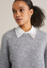 Szary sweter z dzianiny o okrągłym dekolcie, z białym koronkowym kołnierzykiem z misternymi wzorami. Widoczne kręcone włosy i kolczyki wkrętki.