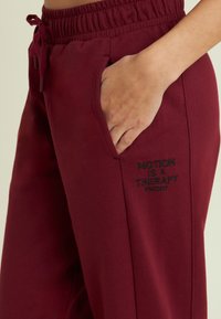 Pantaloni della tuta bordeaux con vita elastica e cordoncino, dotati di tasche laterali e testo ricamato in nero che recita "IL MOVIMENTO È UNA TERAPIA."
