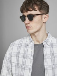 Ochelari de soare cu lentile negre într-o ramă cu model țestoasă. Modelul poartă un tricou gri sub o cămașă albă cu carouri.